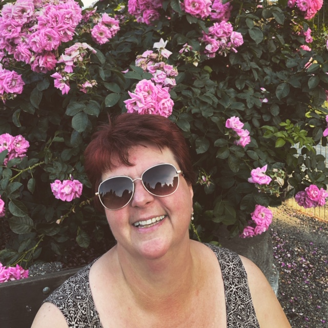 A picture of Ildiko Kalman-Forbes, wearing sunglasses and a big smile as she is sitting on a bench in front of a  gorgeous pink rose bush.